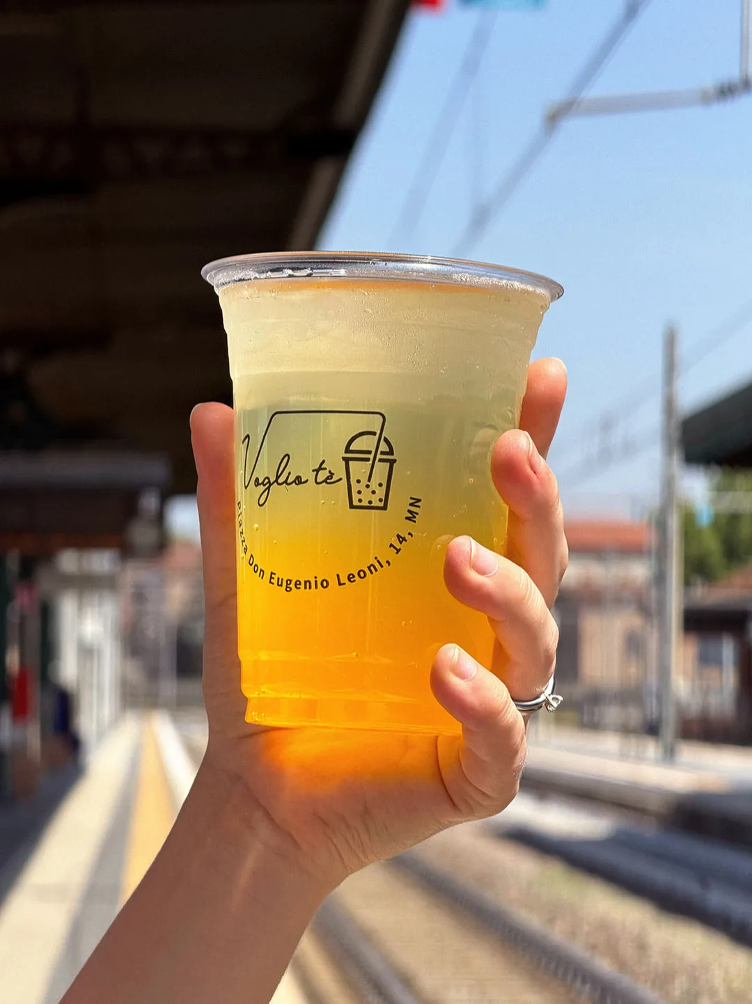 Lemon fruit soda held up on the platform at Mantua station, with the tracks and overhead lines behind it